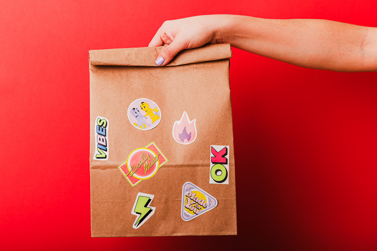Hand holding a brown paper delivery bag with colorful stickers against a red background